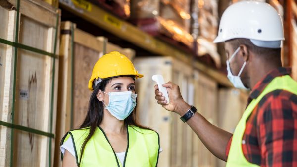 Workers with masks and hard hats getting temperature checked arriving in a warehouse